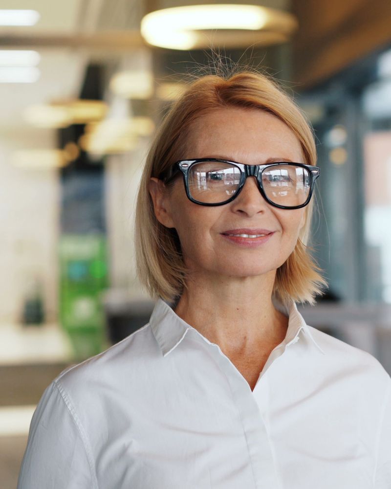 Kobieta w okularach i białej koszuli uśmiecha się w jasnym biurze